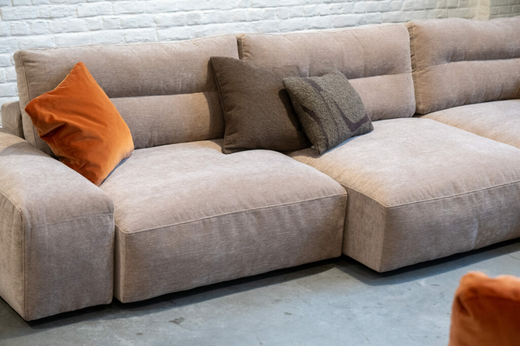 Une composition Expo Canape My Taos en beige, accentuée d'un coussin orange et marron foncé, est placée contre un mur de briques blanches sur le sol en béton, créant un espace accueillant et élégant.