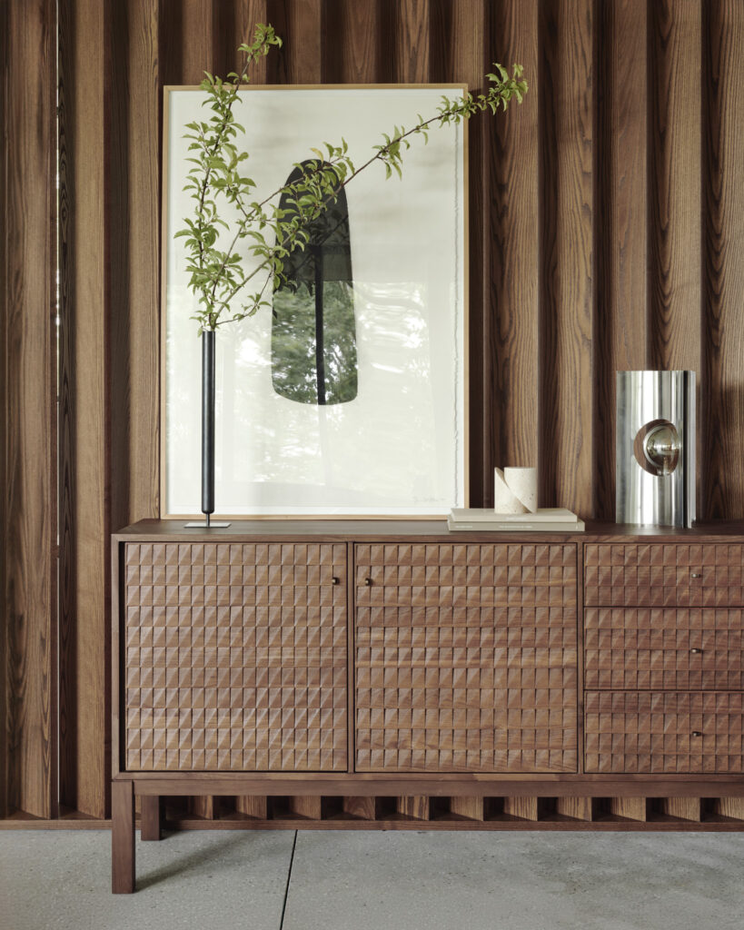Un buffet Sono en bois au motif texturé se dresse contre un mur en bois assorti. Un vase avec des branches vertes, une œuvre d'art abstraite encadrée et un objet de décoration en métal sont placés sur le dessus du buffet Sono.