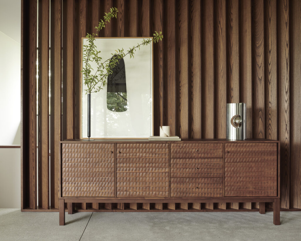 Un buffet en bois à la texture tissée, surmonté d'une œuvre d'art abstraite encadrée et d'une haute branche dans un vase, se dresse contre un mur de lattes de bois vertical, ajoutant aux dimensions élégantes de la pièce.