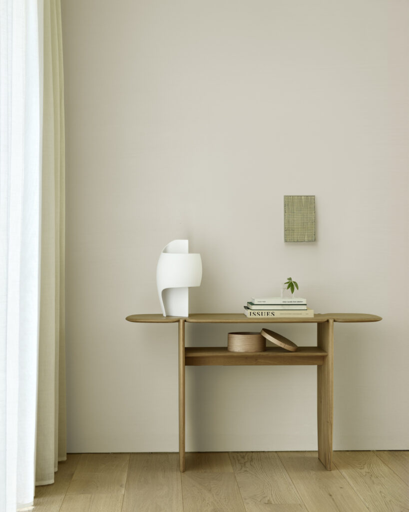 Une console minimaliste en bois Chêne huilé, H : 75, contre un mur de couleur claire, avec une lampe moderne blanche, une pile de livres, une plante et une boîte fermée disposées dessus. Une petite œuvre d'art texturée est suspendue au-dessus.