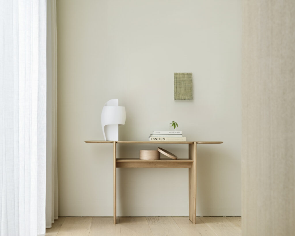 Table minimaliste en bois finition Chêne huilé avec une sculpture blanche, des livres empilés, une petite plante et un récipient en bois contre un mur de couleur claire. Une pièce décorative tissée est suspendue au-dessus de la table. Les dimensions sont parfaites pour n'importe quel espace.