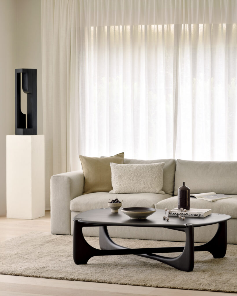 Un canapé beige trône devant des rideaux transparents, accompagné d'une table basse sculpturale PI avec des objets décoratifs. Une sculpture noire moderne sur un piédestal blanc se trouve à gauche.