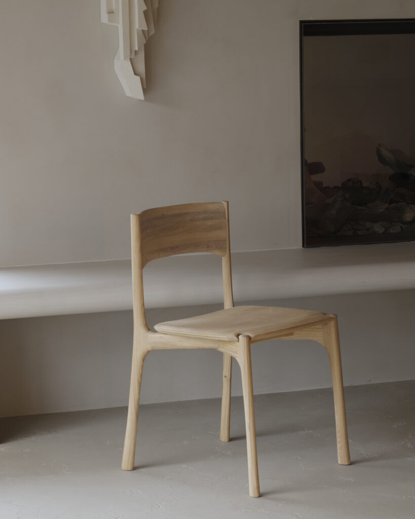 Une chaise de salle à manger PI en bois minimaliste avec un dossier et un siège incurvés est placée dans une pièce de couleur neutre près d'une décoration murale et d'une cheminée vide.