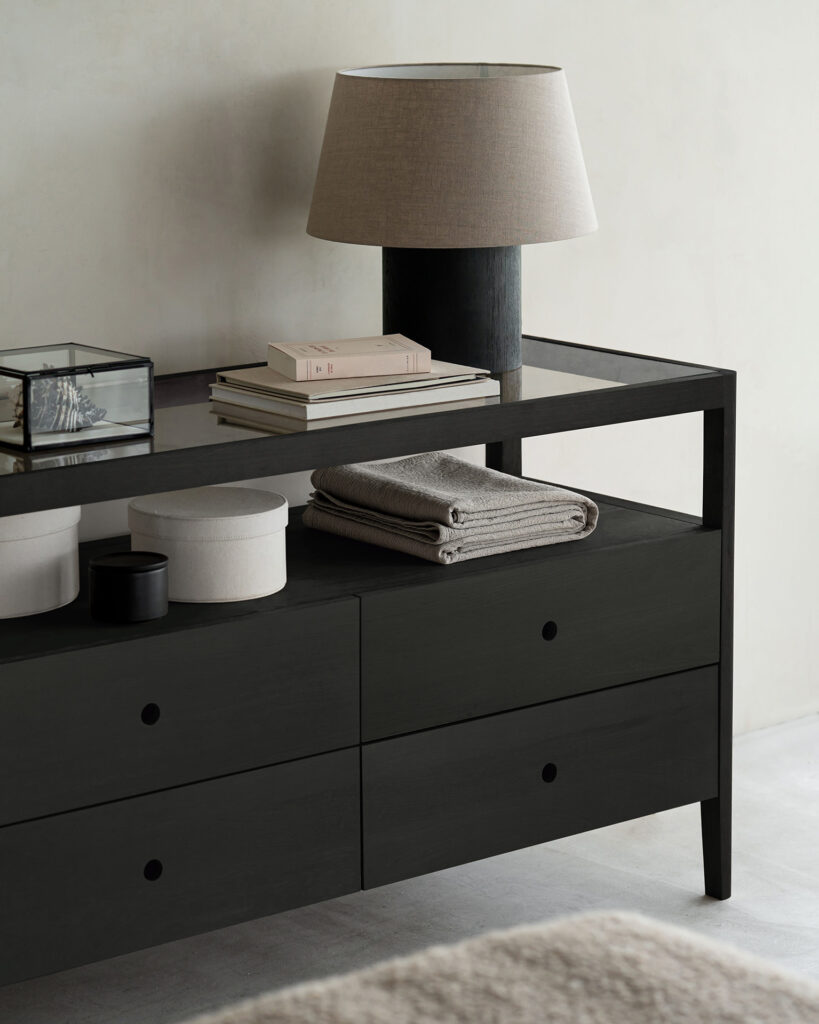 Une table d'appoint en bois foncé, rappelant une commode classique en chêne, supporte une lampe de table, une boîte en verre et quelques livres sur le dessus. Des vêtements pliés et des récipients ronds sont placés sur une étagère inférieure.