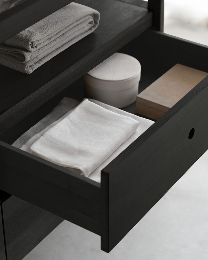 Un tiroir partiellement ouvert dans la commode en chêne révèle une serviette blanche soigneusement pliée, un petit récipient blanc, une boîte beige et du linge blanc plié supplémentaire sur l'étagère au-dessus.