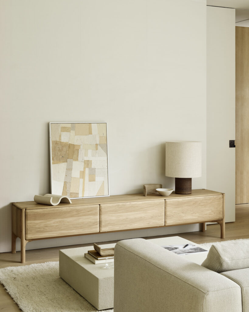 Un salon minimaliste comprend un meuble TV PI en bois clair de Chêne huilé avec une œuvre d'art géométrique, une lampe et un bol. Un canapé beige et un tapis texturé sont au premier plan. Les dimensions précises du meuble offrent à la fois fonctionnalité et style.