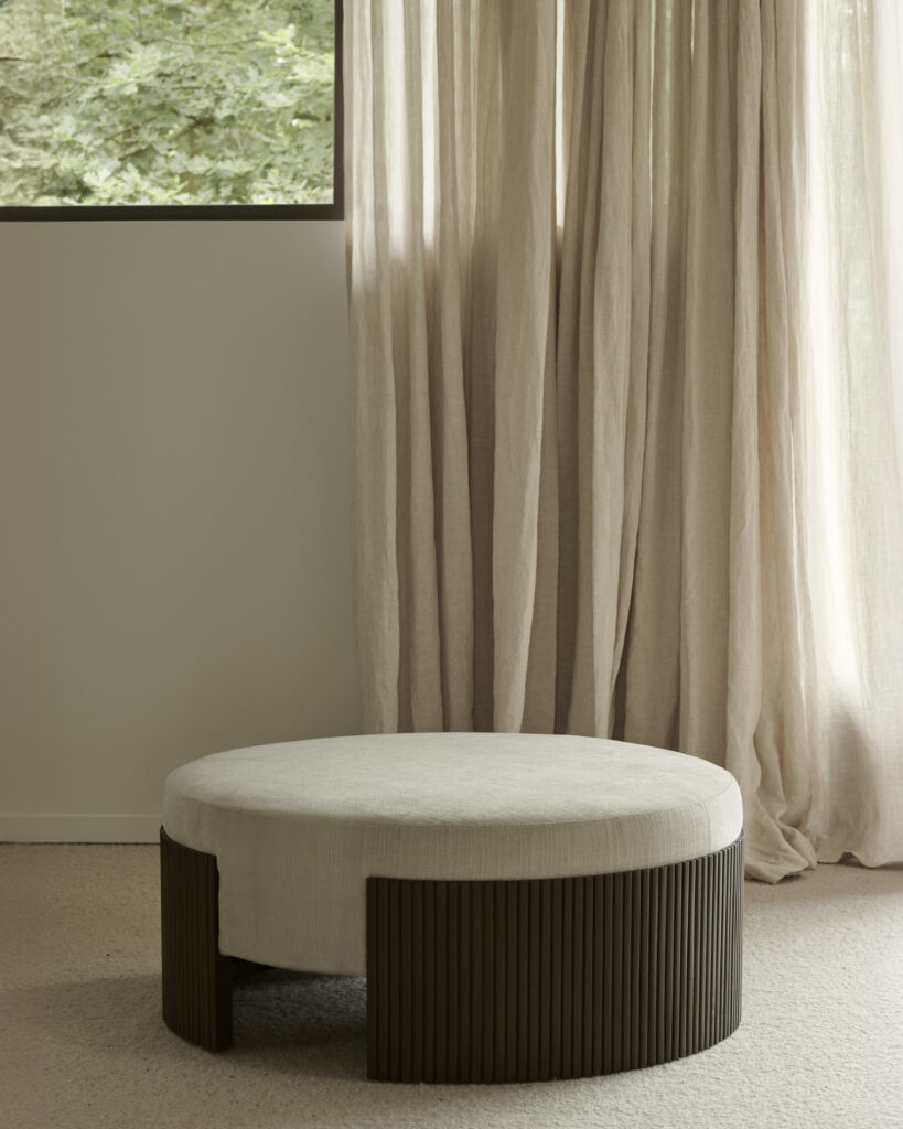 Le Pouf Roller Max, aux dimensions de 85x85x35, est un pouf rond caractérisé par un plateau beige et des pieds cylindriques foncés, parfaitement placé devant une fenêtre ornée de rideaux beige clair.
