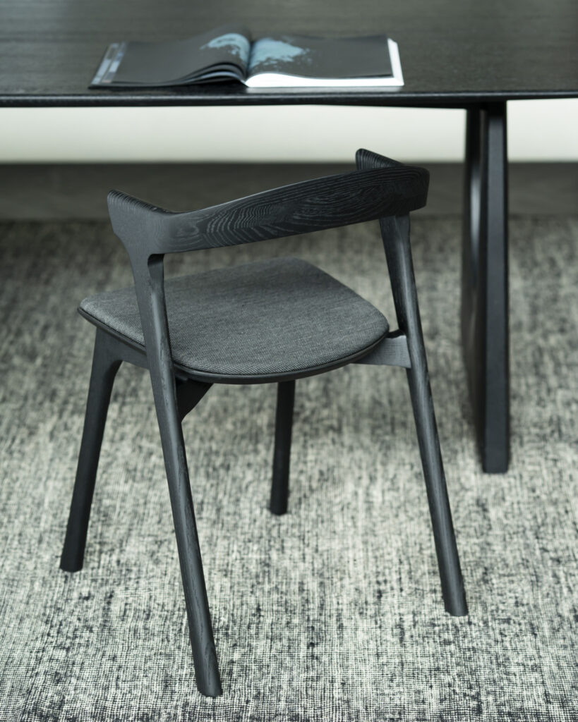 Une chaise Bok en bois noir avec un siège rembourré gris est placée devant une table noire sur un tapis gris texturé. Un livre ouvert est posé sur la table.