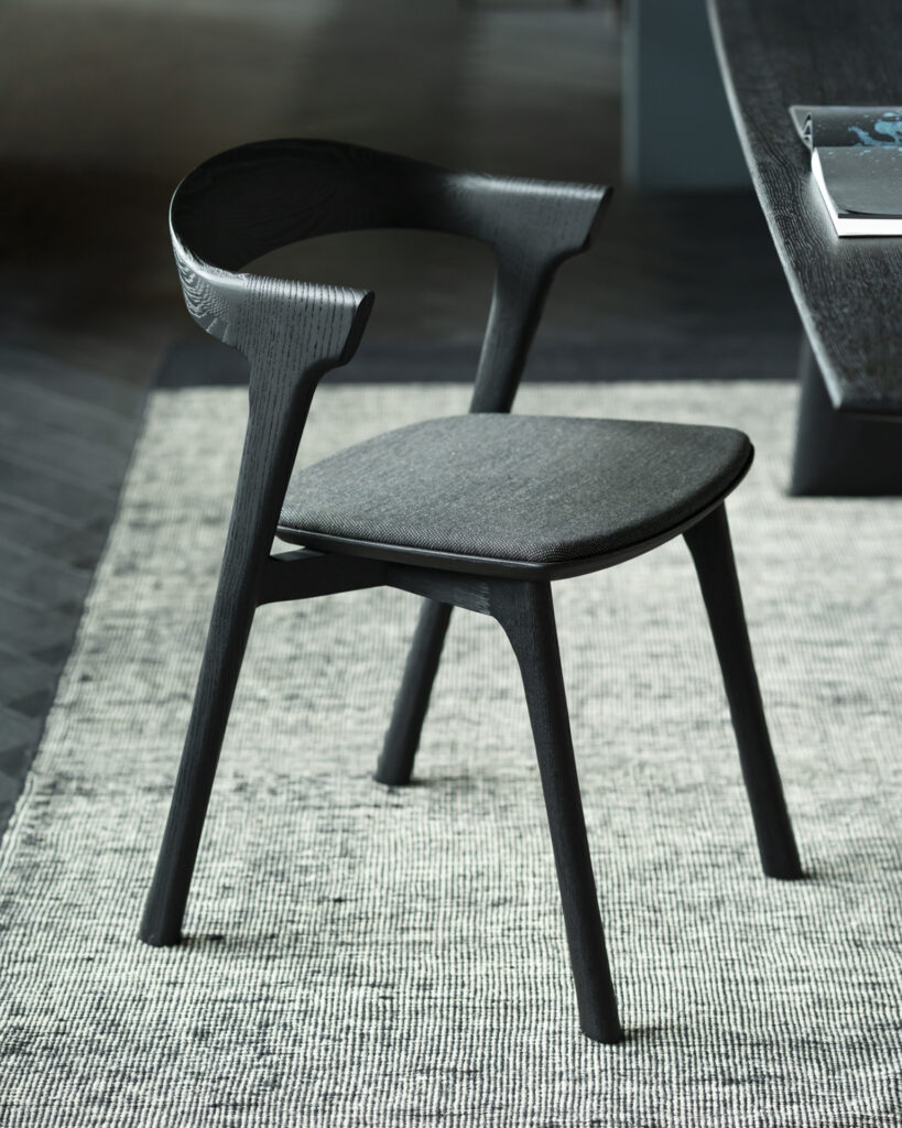 Une chaise moderne en bois de chêne noir avec un dossier incurvé et un siège rembourré en tissu gris posé sur un tapis texturé à côté d'une table.