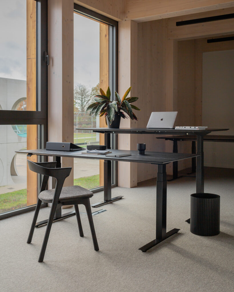 Un bureau à domicile moderne avec un bureau noir, un ordinateur portable, une tablette, une chaise Bok avec revêtement en tissu gris et une plante en pot près de grandes fenêtres offrant une vue sur un espace extérieur verdoyant.