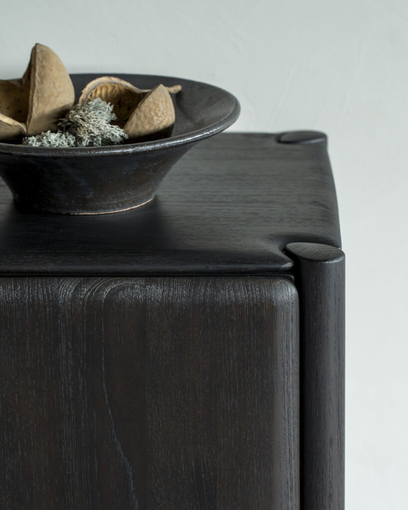 Le Buffet PI en teck 2 portes, fabriqué en bois de teck foncé aux coins arrondis et orné d'un bol foncé sophistiqué rempli de plantes séchées, offre une esthétique de buffet élégante.