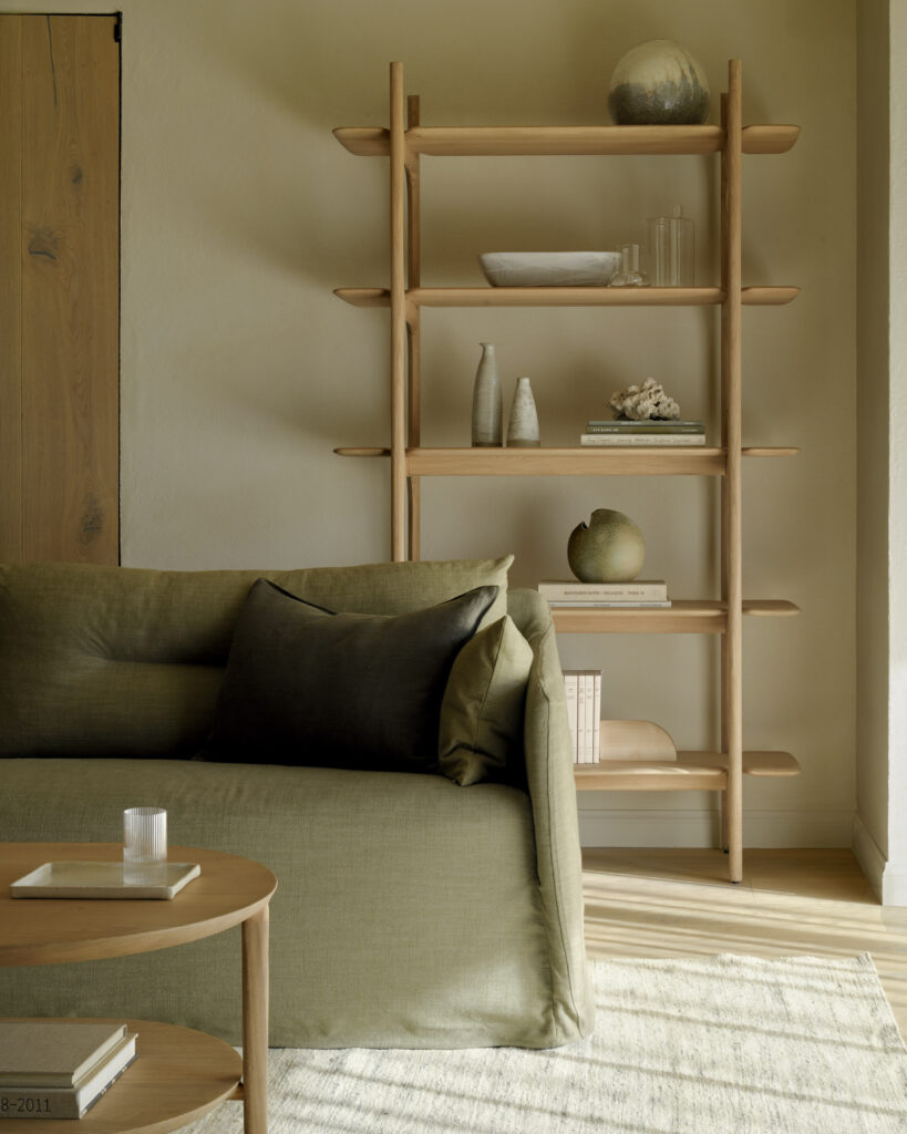 Un salon moderne avec un canapé vert, une table basse ronde en bois et l'étagère PI présentant des vases, des bols et des livres, le tout baigné de lumière naturelle.