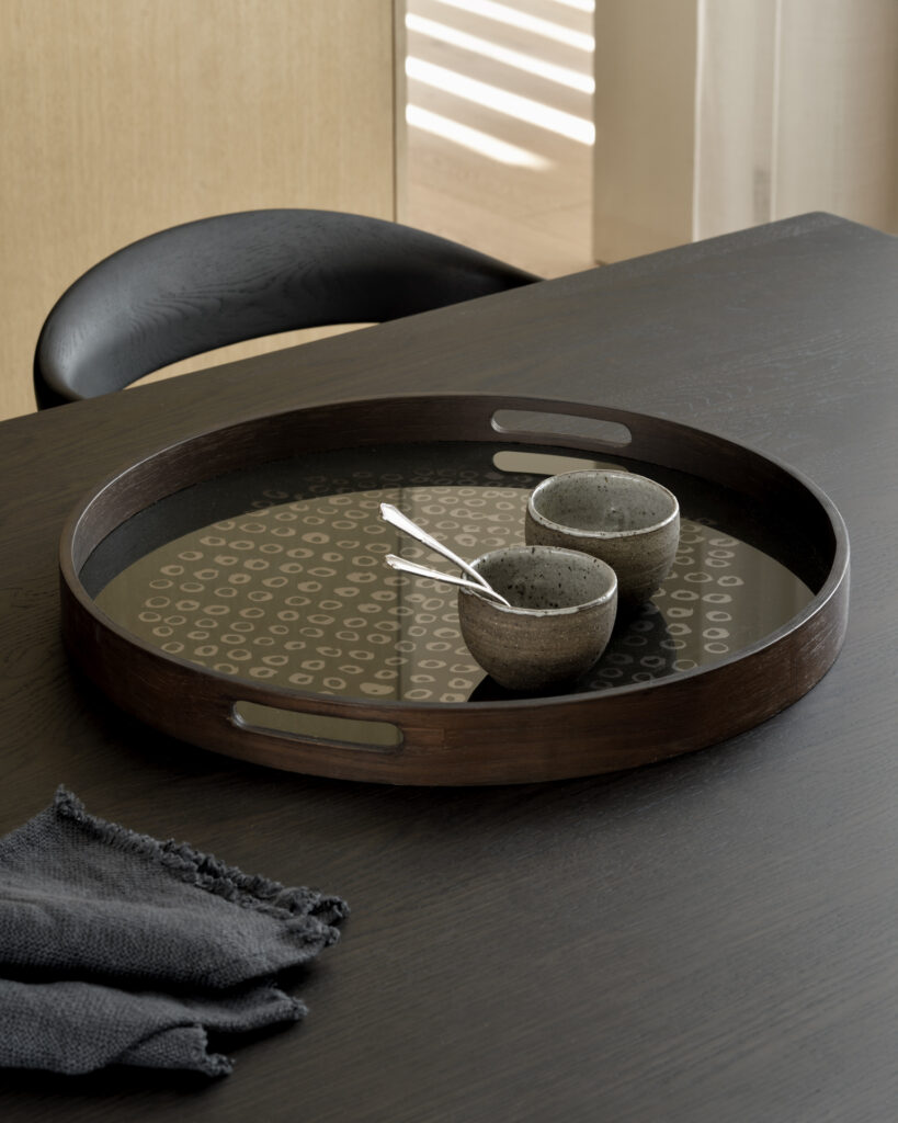 Un plateau rond en verre Seconde Nature Moss avec deux petites tasses en céramique et une cuillère en métal repose sur une table sombre, accompagné d'une serviette en tissu foncé pliée, ajoutant une touche d'élégance au cadre serein.