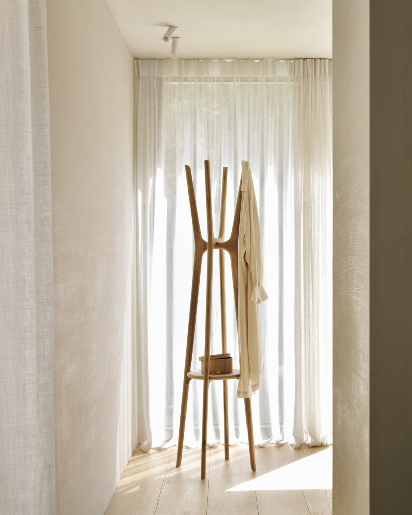 Chambre minimaliste avec un portemanteau en chêne naturel sur lequel sont posés vêtements et livres. Une douce lumière naturelle filtre à travers des rideaux blancs transparents, illuminant le parquet en bois clair.