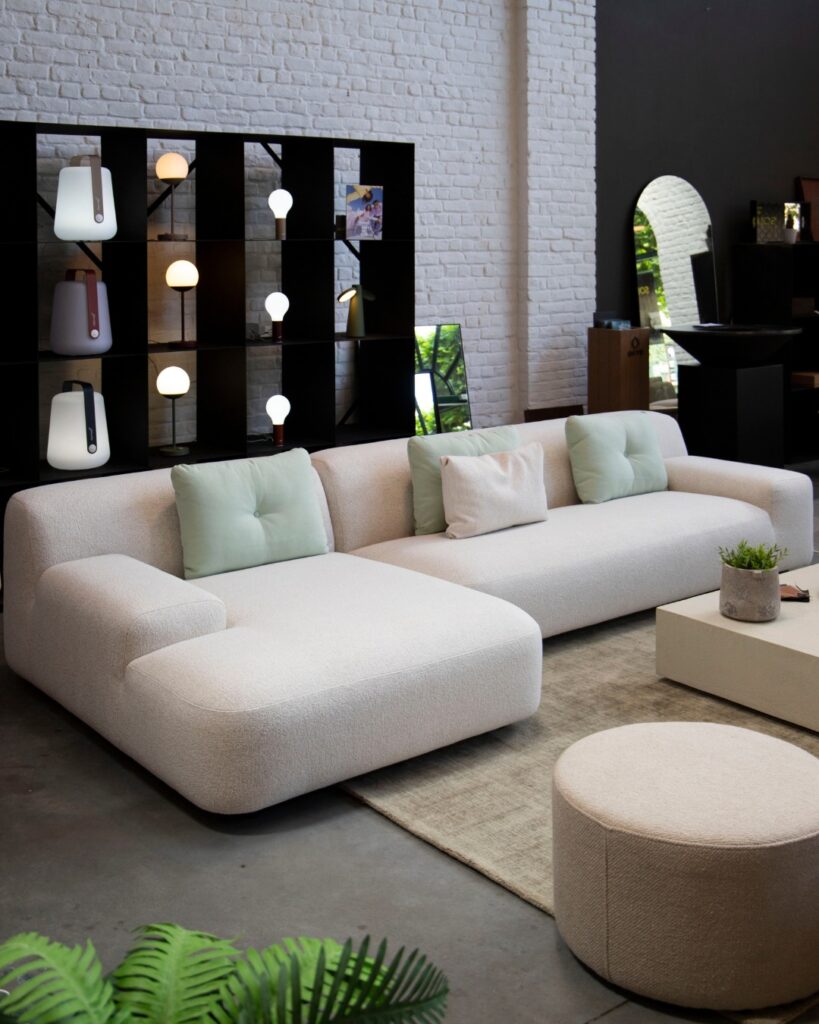 Salon moderne avec un Canapé Jason blanc, tissu 1434 en forme de L, des coussins clairs, un pouf rond, une plante en pot, une étagère avec lumières et un grand miroir contre un mur de briques.