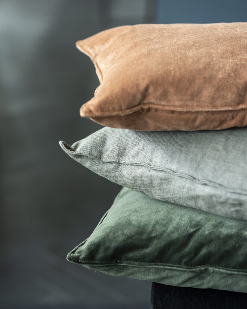 Un trio de coussins en velours sont empilés, présentant un coussin marron clair sur le dessus, un coussin gris au milieu et un coussin vert foncé en bas, le tout sur un fond flou.
