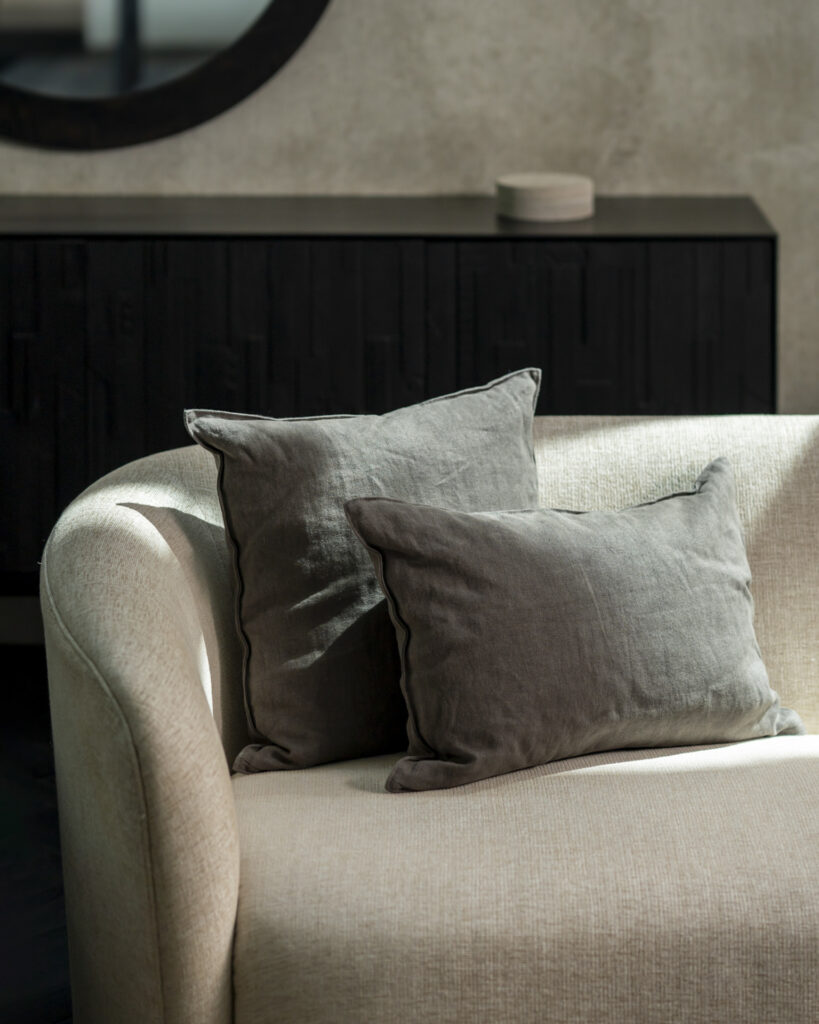Un canapé beige orné de deux coussins gris foncé et d'un élégant coussin Lin est placé devant un meuble noir épuré. La scène est magnifiquement éclairée par la lumière naturelle.