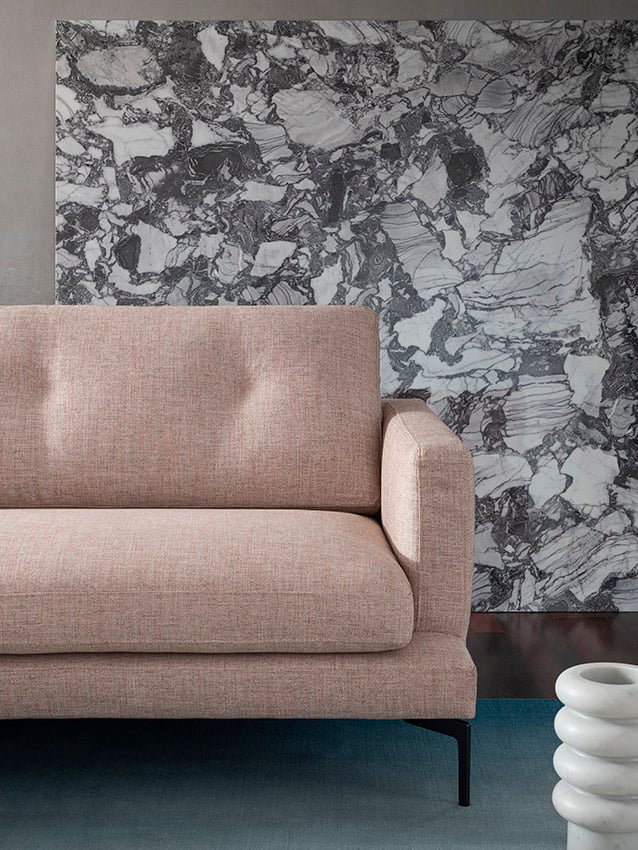 Un Expo-Canapé l'Essentiel 220 A en rose clair est placé devant un mur de marbre gris et blanc, avec une table d'appoint sculpturale blanche partiellement visible sur la droite.