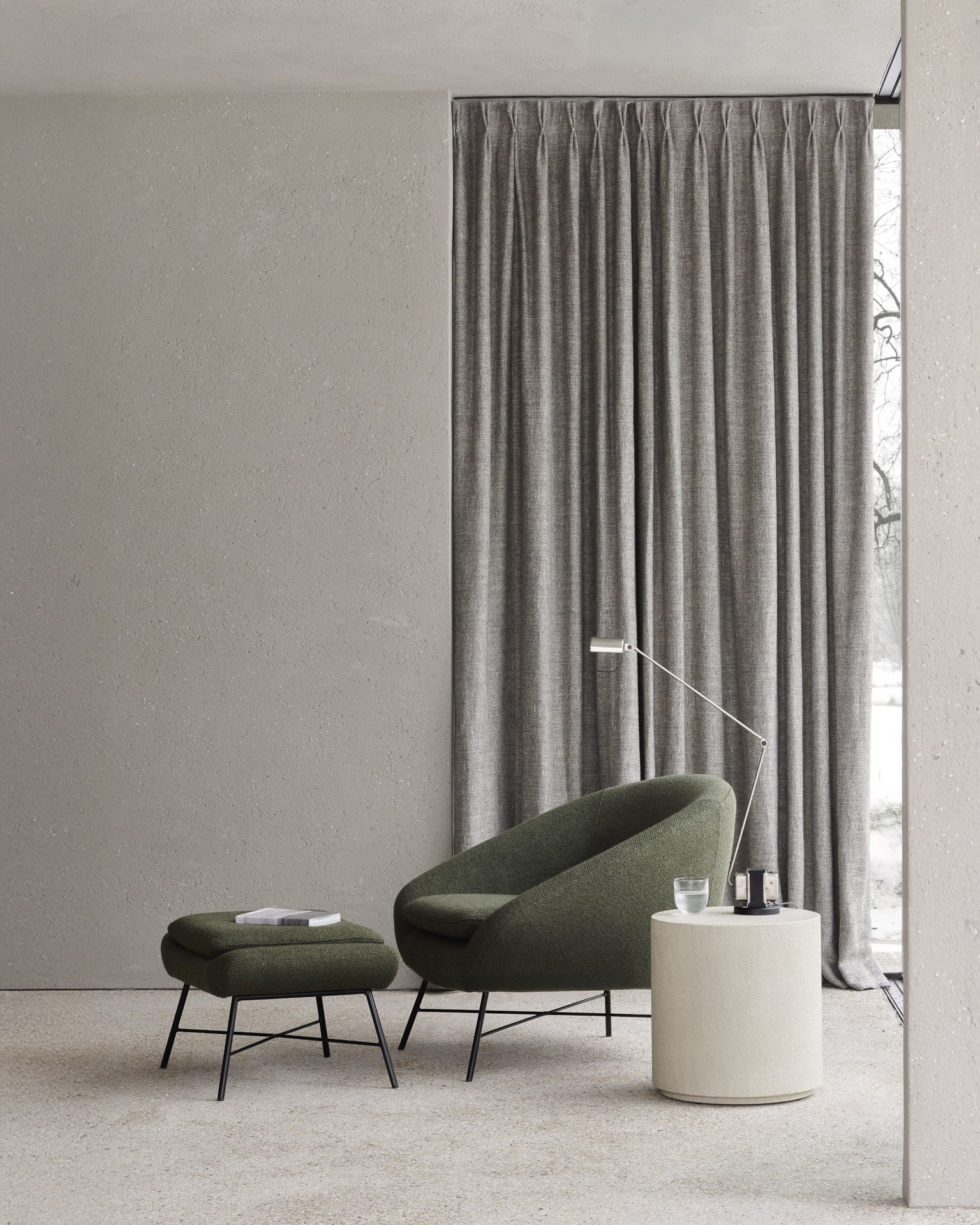 Un espace de vie minimaliste avec un fauteuil vert, le pouf Barrow Repose-pieds, une table d'appoint ronde contenant un verre et un livre, un lampadaire, des rideaux beiges et une grande fenêtre.