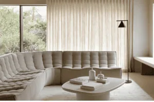 Salon minimaliste avec canapé sectionnel incurvé gris, table basse blanche, lampe et grande fenêtre avec rideaux fermés. Palette de couleurs neutres rehaussée par la lumière naturelle entrant par la fenêtre.