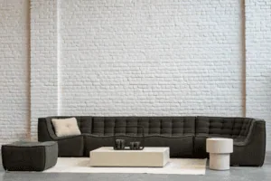 Un canapé sectionnel gris avec un coussin blanc est centré sur un tapis blanc. Une table basse carrée et une petite table d'appoint ronde complètent le décor contre un élégant mur de briques blanches.