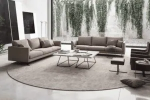 Salon moderne comprenant deux canapés gris, un tapis rond et un mur de verre offrant une vue sur un jardin aux hauts murs recouverts de lierre.