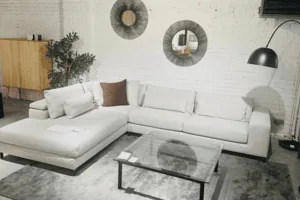 Salon moderne avec canapé d'angle blanc, lampadaire, deux miroirs muraux ronds, tapis gris et table basse à plateau en verre. Une armoire en bois et une plante en pot ornent gracieusement l'arrière-plan.