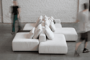 Deux silhouettes floues passent devant un canapé modulaire blanc moderne orné de coussins, posé contre un mur de briques blanches immaculées.