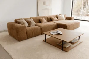 Un canapé beige avec plusieurs coussins est placé dans un salon minimaliste. Il repose sur un tapis neutre à côté d'une table basse en bois sur laquelle est posé un magazine. De grandes fenêtres et des œuvres d'art murales abstraites complètent cet espace serein.