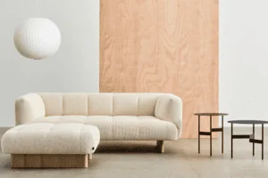 Un salon minimaliste comprenant un canapé beige, un pouf, deux tables rondes en bois, une lanterne en papier et des boiseries claires sur le mur.