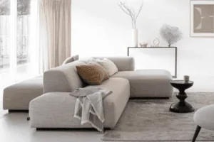 Salon minimaliste avec canapé d'angle beige, couverture drapée, petite table ronde et éléments décoratifs sur une console. Palette de couleurs neutres avec lumière naturelle.