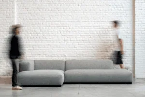 Deux silhouettes floues passant devant un canapé gris contre un mur de briques blanches.
