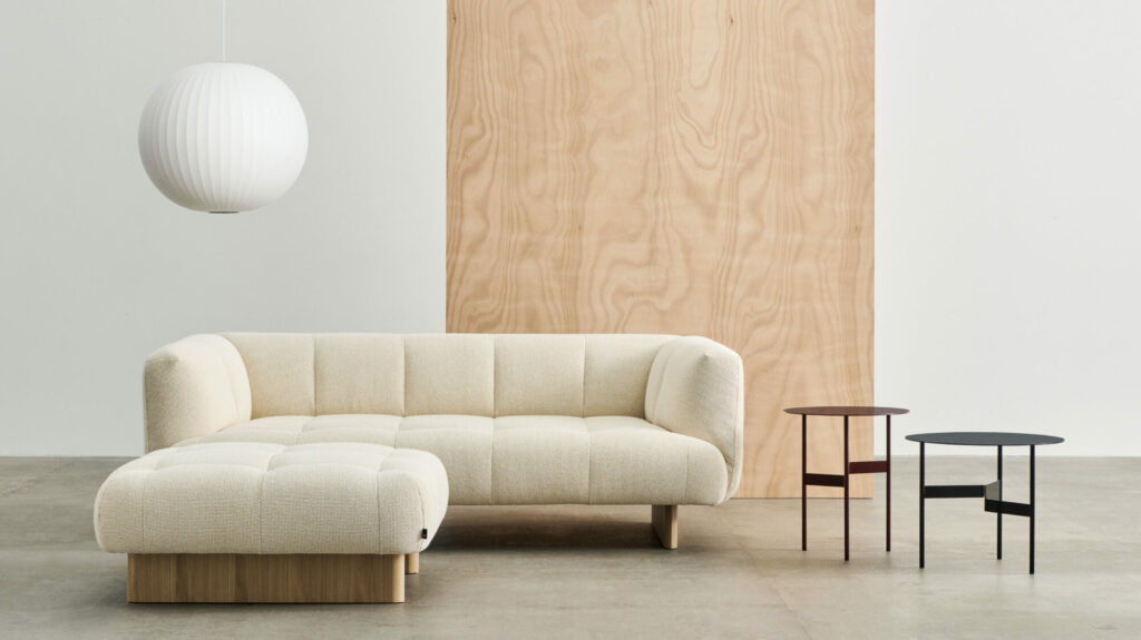 Un salon minimaliste comprenant un canapé 2 places Quilton Lift avec une base en chêne laqué en Tartaglia Ocre White, accompagné de deux tables d'appoint rondes noires, d'une grande suspension et d'un fond en panneaux de bois.