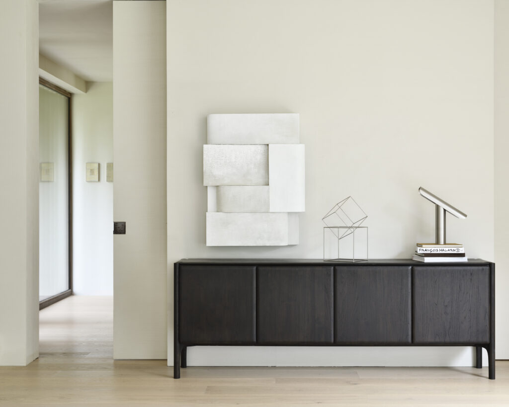 Un intérieur minimaliste comprenant un élégant buffet en teck foncé de Buffet PI, teck brun foncé, 4 portes, complété par une sculpture géométrique, des livres, une lampe moderne et une œuvre d'art murale abstraite.