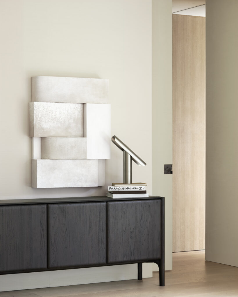 Une pièce minimaliste présente une sculpture moderne au-dessus d'un buffet en bois sombre, ressemblant à l'élégant Buffet PI en teck foncé à quatre portes. Une lampe de bureau en métal repose sur des livres empilés, ajoutant une touche intellectuelle.