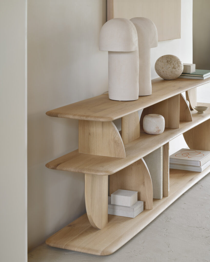 Une étagère en bois, fabriquée en chêne naturel et rappelant une console géométrique, accueille des sculptures abstraites, des livres empilés et des objets décoratifs dans une pièce minimaliste. Sa finition huilée rehausse la simplicité et l'élégance de l'espace.