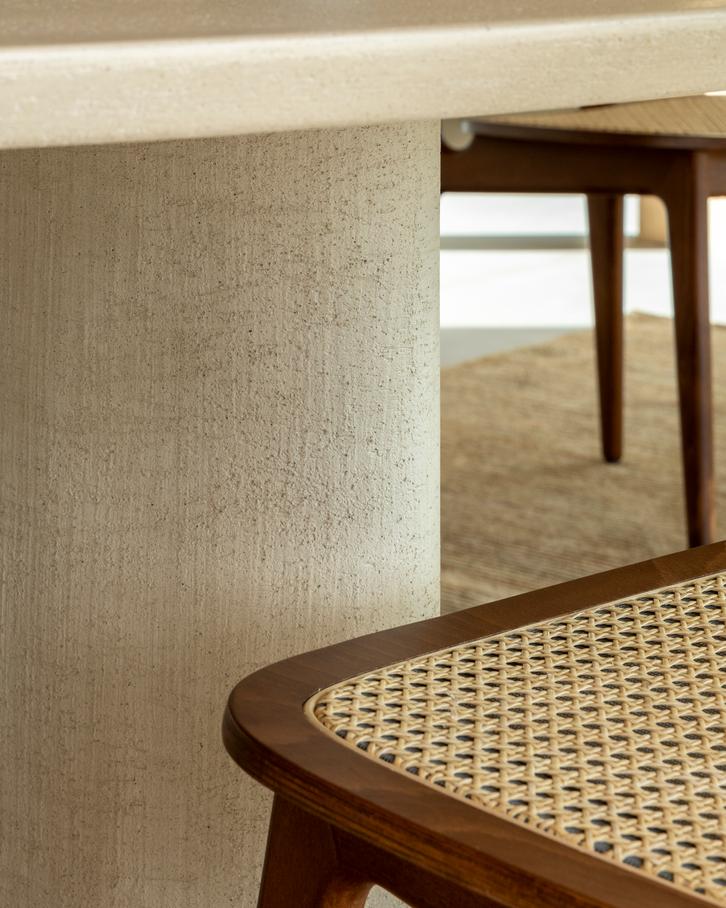 Gros plan sur la table ronde texturée Table ovale Ari et sur une chaise en bois avec assise en rotin tressé, posées sur un tapis neutre.