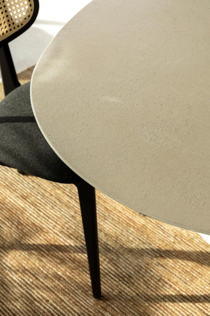 La Table ovale Ari en beige avec une surface lisse est associée à une chaise noire avec un dossier en rotin, posée sur un tapis tissé rayé.