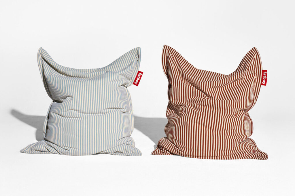 Deux poufs rayés, l'un en bleu Blizzard et blanc, l'autre en crème Cognac et blanc, avec des étiquettes de marque rouges, sont placés côte à côte sur un fond blanc immaculé.