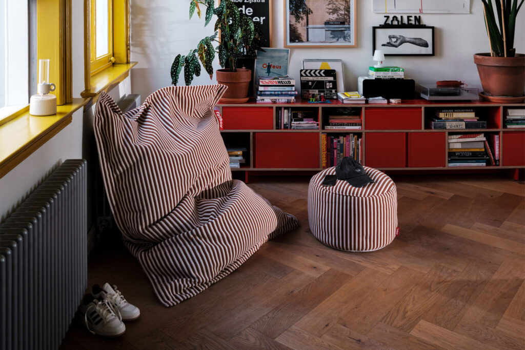 Salon cosy avec poufs rayés et armoire rouge remplie de livres. Des plantes en pot se mêlent aux œuvres d'art murales, tandis qu'une fenêtre aux bordures bleu Blizzard illumine l'espace. Des baskets reposent sur le parquet près d'un radiateur, ajoutant une touche de charme au décor.