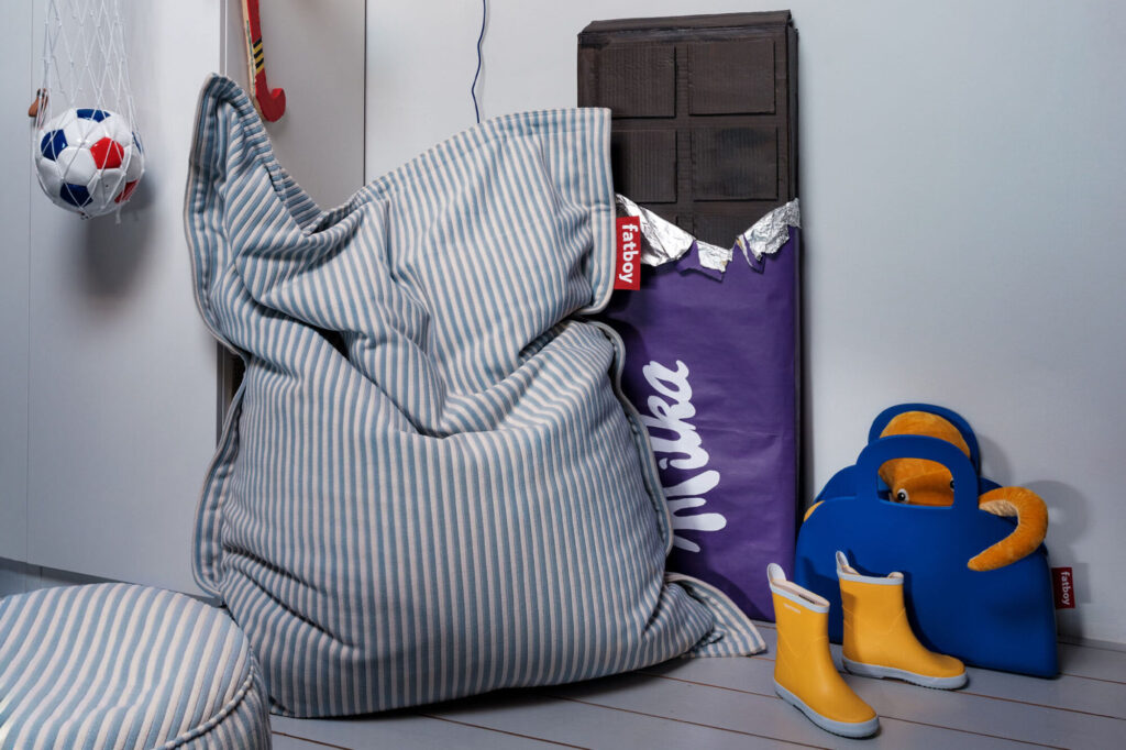 Une pièce avec un pouf rayé, une décoration de barre chocolatée Cognac Creme via Original Slim Terry, des bottes jaunes, un ours en peluche dans une chaise bleue et un filet de ballon de football suspendu.