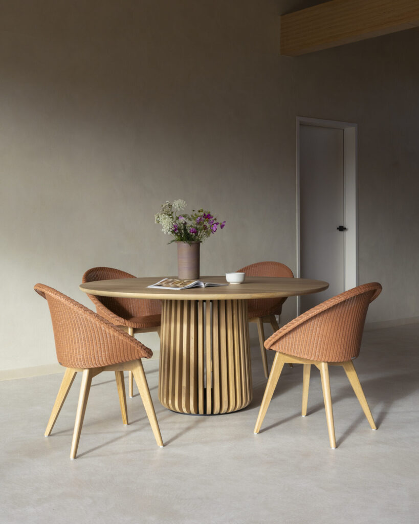 Une table ronde Maru avec un vase de fleurs et un magazine est entourée de quatre chaises en osier dans une pièce minimaliste aux murs beiges et une porte blanche fermée.