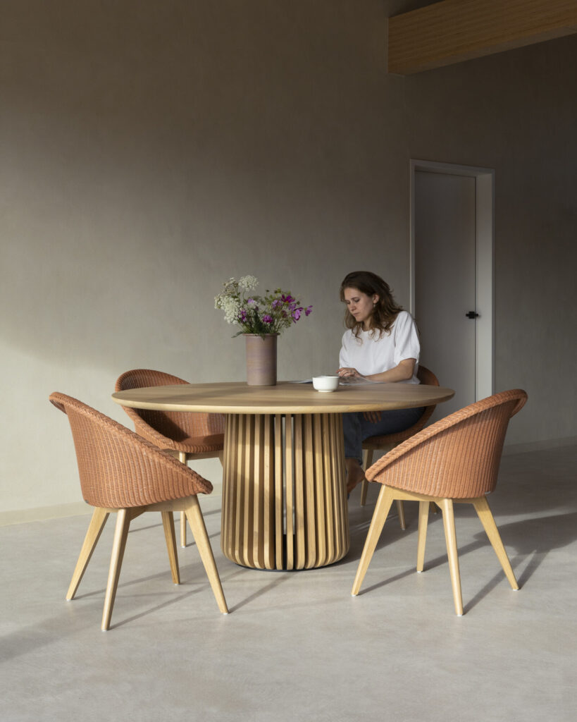 Une personne est assise à une table ronde Maru avec un vase de fleurs et une tasse, entourée de trois chaises tissées dans la pièce minimaliste.
