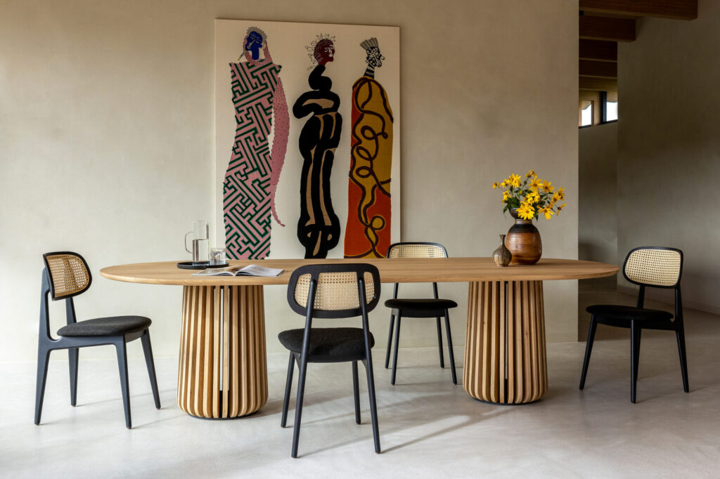 Une salle à manger moderne présente une table Maru - ovale, entourée de cinq chaises. Un vase de fleurs jaunes au centre met en valeur l'art abstrait sur le mur.