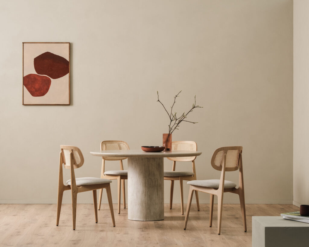 Une salle à manger minimaliste est dotée de la table Ari - ronde, entourée de quatre élégantes chaises en bois. Un vase de branches trône au centre de la table, tandis qu'une œuvre d'art géométrique rouge anime le mur beige, complétant parfaitement cet ensemble de meubles harmonieux.