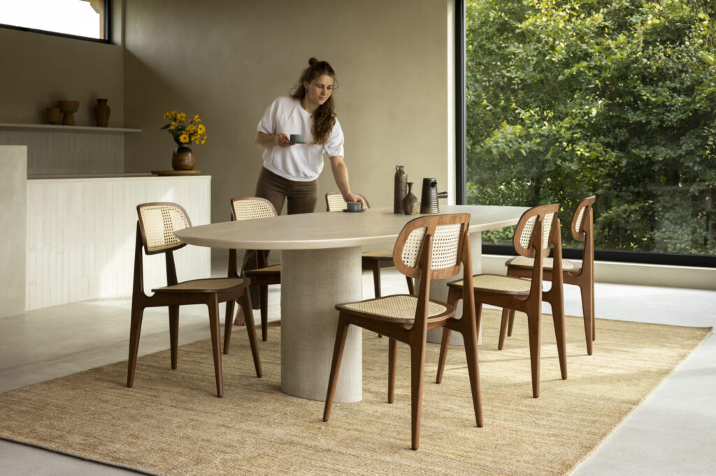 Une personne installant une élégante table Ari dans une pièce minimaliste avec des chaises en bois, sur fond d'une grande fenêtre donnant sur la verdure.