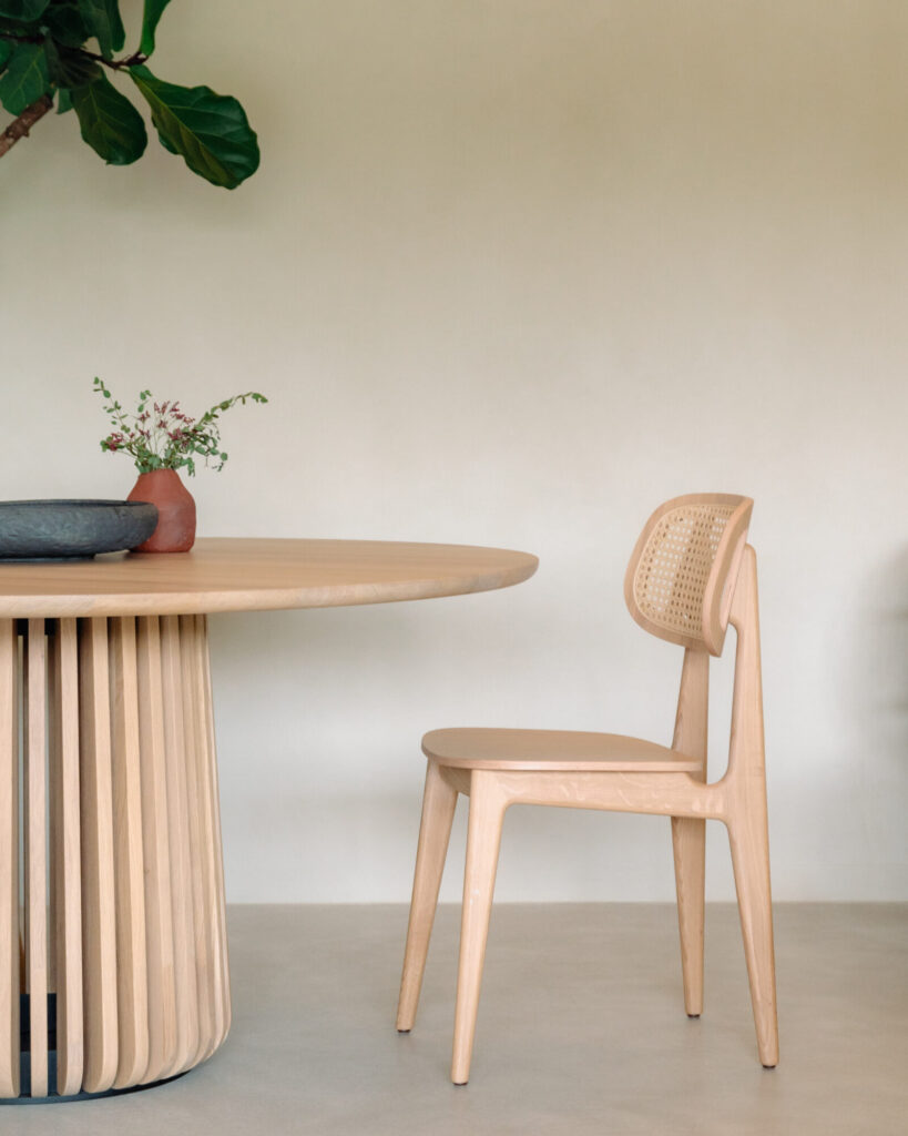 Une chaise en bois au dossier tressé, rappelant la Chaise Titus, trône à côté d'une table ronde en chêne naturel. Une petite plante dans un pot en terre cuite et un plat gris ornent le plateau de la table.