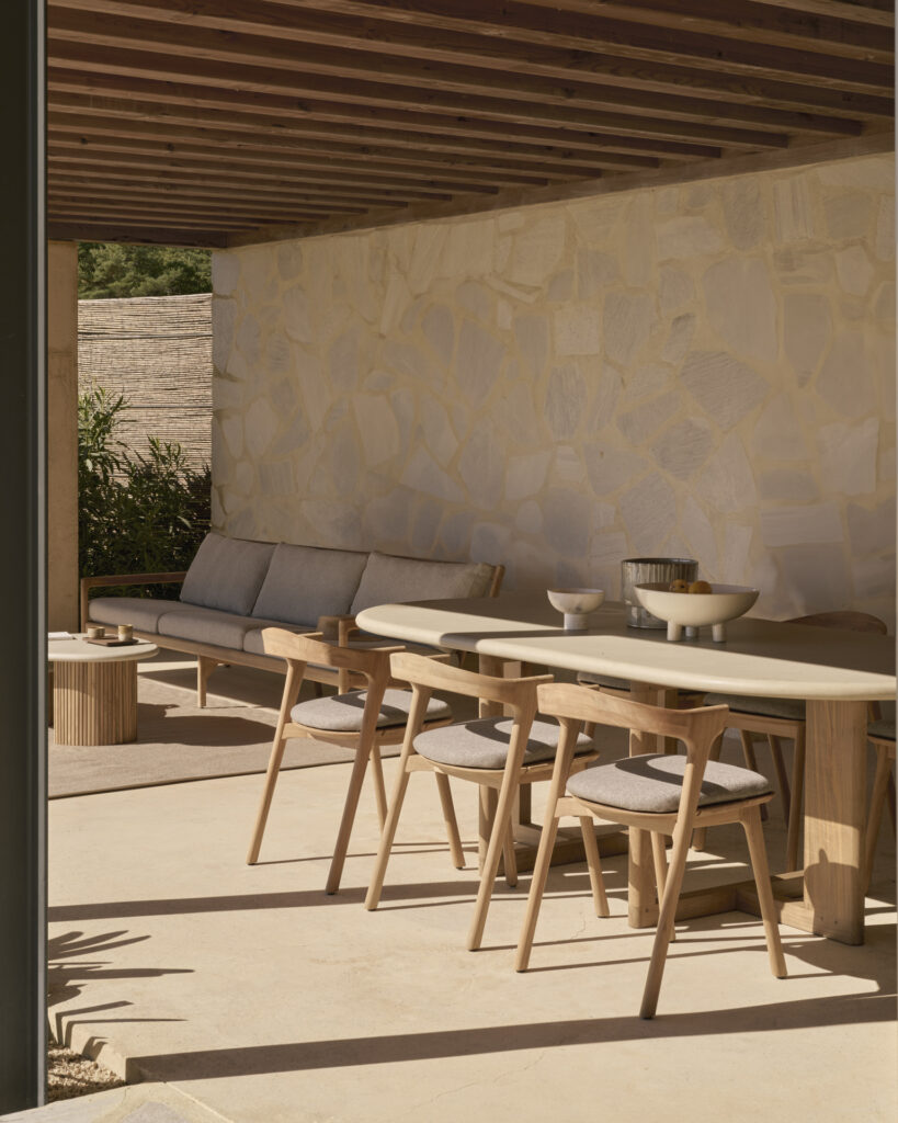La terrasse extérieure est dotée d'un mur en pierre et d'un plafond en bois, avec des meubles comprenant un canapé rembourré, des chaises, une table à manger extérieure à tréteaux et une petite table décorative. Une plante en pot ajoute du charme au décor.