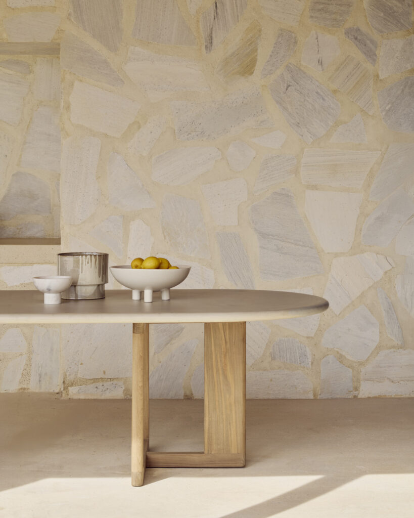 Une table à manger d'extérieur à tréteaux avec une base en bois clair comporte un bol de citrons jaunes, un petit bol et un récipient métallique contre un mur carrelé en pierre.