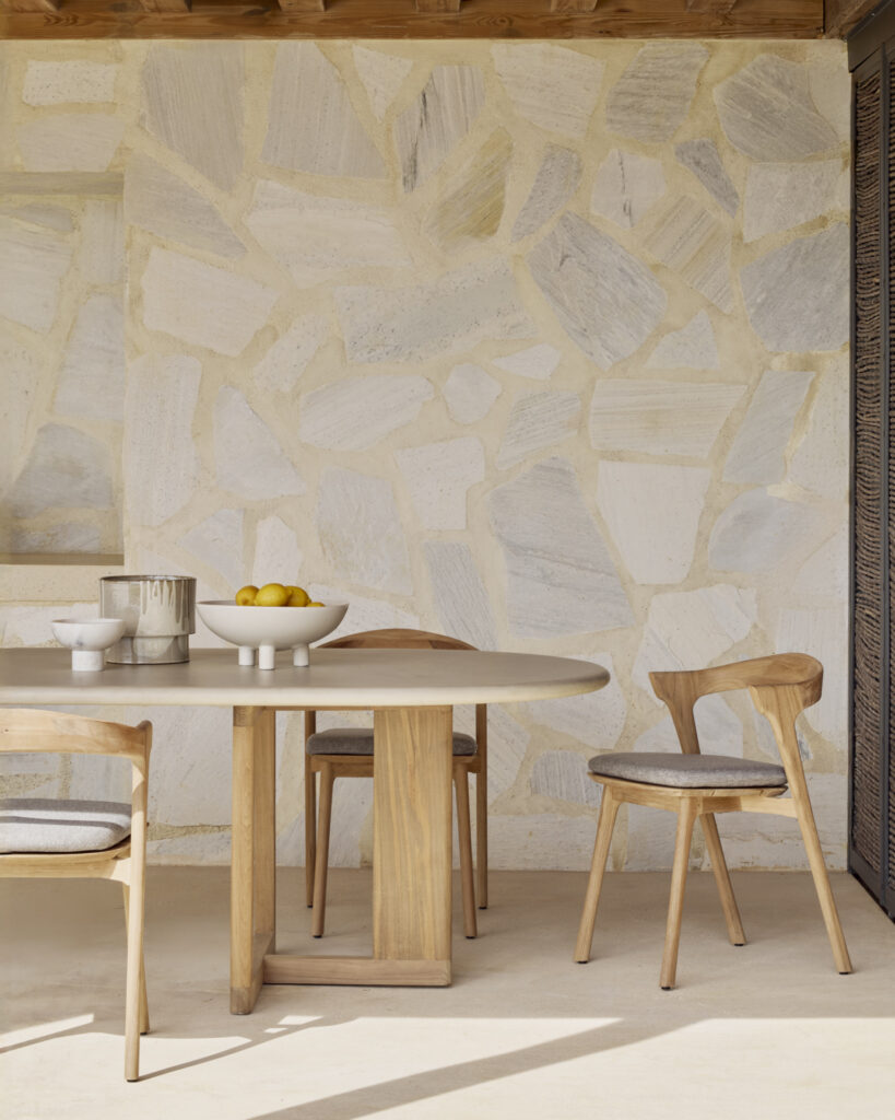 Une table à manger d'extérieur à tréteaux et des chaises rehaussent un patio près d'un mur de pierre texturé, avec un bol de citrons et un pichet en métal ajoutant du charme.